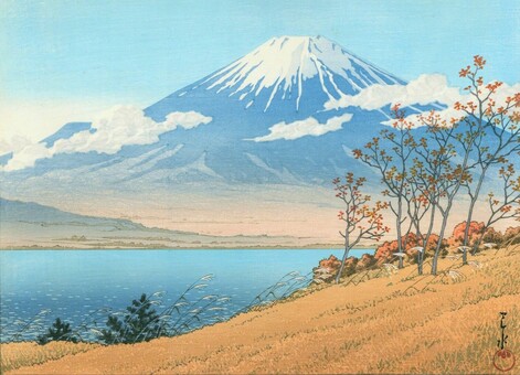 A view over Lake Yamanaka looking toward Mount Fuji on a sunny autumn day. In the foreground, the grass is brown and the thinning leaves on the trees are orange as autumn takes hold; trees and taller grasses are pushed to the right as wind blows in off the lake toward shore. Beyond, the lake is quiet, unoccupied by any boats; on the far side of the water, faint mist obscures details of the gentle hills around the base of snow-capped Mount Fuji, rising against a clear blue sky