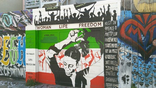 Photo d'un graff dans une ruelle de San Francisco. En fond, le drapeau iranien horizontal vert blanc rouge. Par dessus, une femme en noir et blanc se coupe sa queue de cheval. En haut est noté WOMAN LIFE FREEDOM.