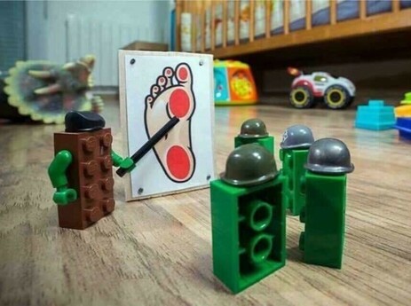 A Lego in a helmut to the left, pointing to a board in center of frame with a picture of a foot with two "strike zones" in red, with several helmeted Legos to the right paying careful attention. Because we all need a laugh today. 💞