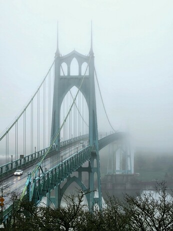 The St Johns Bridge, a suspension bridge painted greenish blue, like the patina on the Statue of Liberty. The towers have cutouts that look like the gothic pointed arches of medieval cathedrals. There is a dense fog so only the nearest tower is visible. The other tower and most of the opposite bank is obscured by the fog.
