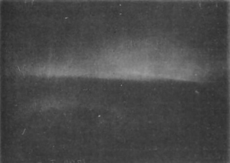 A black and white photo of an aurora, which appears as a nebulous, veil-like cloud in a dark sky.