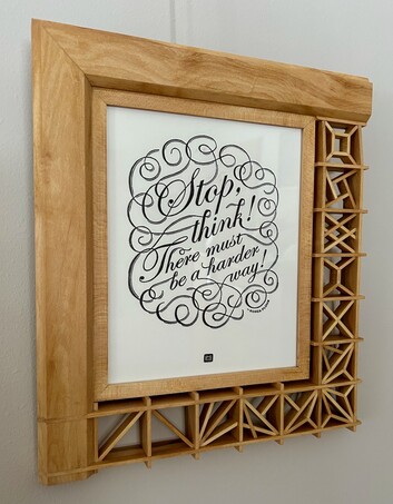 A print reading, "Stop, think! There must be a harder way! ~Roger Plumb" in a wooden frame that is simple solid wood on the top and left edges, and a kumiko lattice on the bottom and right edges.
