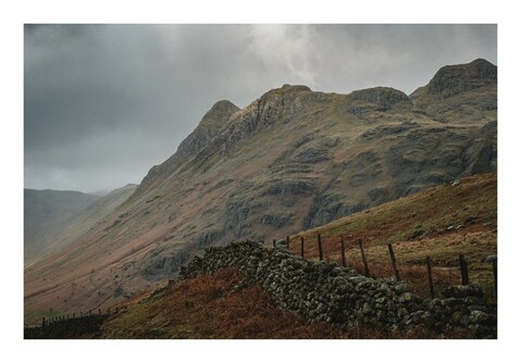 Mountainous landscape with overcast skies, a stone wall, and a wire fence.