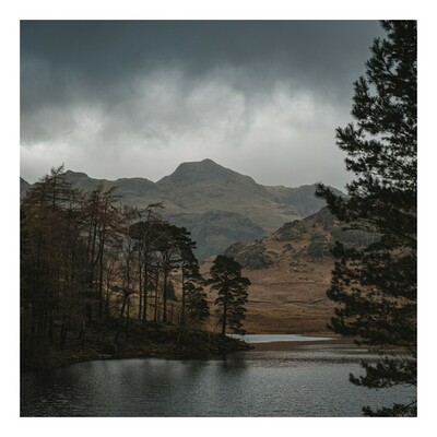 A mountainous landscape with a cloudy sky, a lake in the foreground, and trees on its bank.
