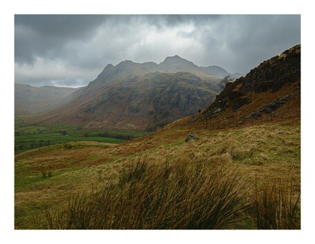 A rugged landscape with grassy fields in the foreground, a fence along the right, and towering, cloud-covered mountains in the background.