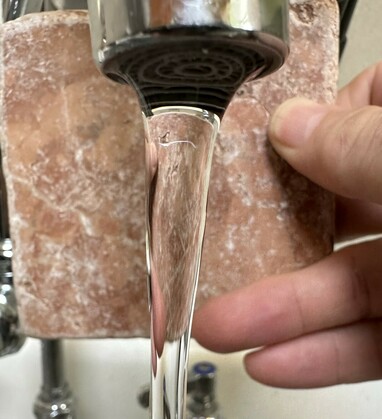 Closeup of laminar flow coming out of a faucet. A hand is holding a stone tile behind the flow to highly the crystalline clarity. The flow looks like a solid bar of glass, as if you could catch a hold of it and hang it on your yule tree.