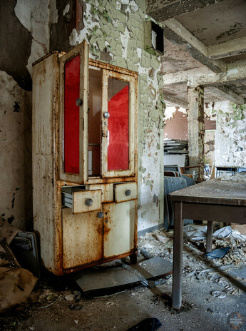 Vertical photo of a rusting metal medicine cabinet. Two of the drawers are open and one of the doors is ajar. The doors have windows with what appears to be red paper over them to obscure what is inside. On the right, a table, desks, and chairs are visible in a decaying room