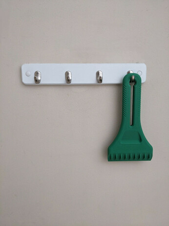 A photo of a dark green, 3D printed ice scraper, hanging on a set of four hooks on a wall.