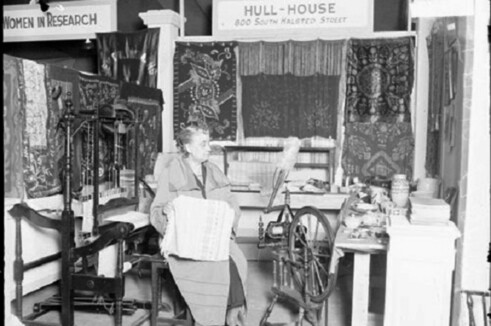B&W photograph. Jane Addams sitting in what appears to be a sewing room at Hull House in 1927. A sign visible on the wall reads "Women In Research."
