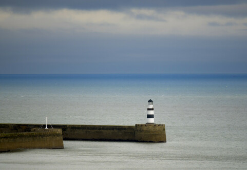 A black and white lighthouse stands at the entrance to a harbour. Behind the sky is dark and moody from a passing rain shower.