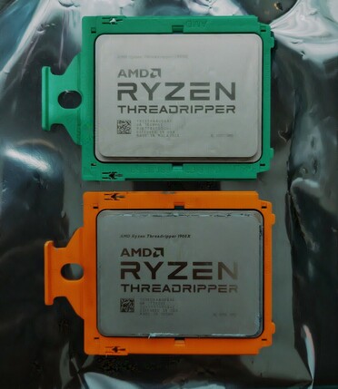 Two AMD Threadripper CPU's placed on a silvery antistatic surface. Top one is a 2950x with a green bracket, bottom one is a 1900x with an orange bracket.