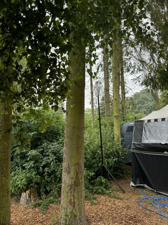 Photo taken of our folding camper which has been pitched up in amongst the trees in a small wood on a camp site. Green ground cover from stinging nettles and wild bushes. A black tripod with a yellow CAT5 cable goes up to a MikroTik LTE based LHGR.