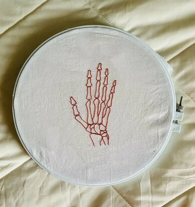 Embroidery of a skeletal hand in red thread on white fabric, held in a circular embroidery hoop.