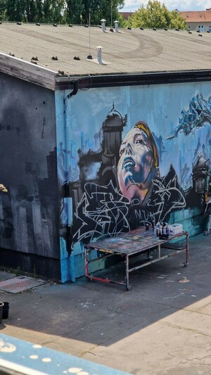 Eine Hauswand mit einem unfertigen Graffiti und einer Bank davor. Auf dem Graffiti ist das Gesicht einer Person zu sehen, im Hintergrund Umrisse einer Skyline. Alles (bisher) in Blautönen gehalten.