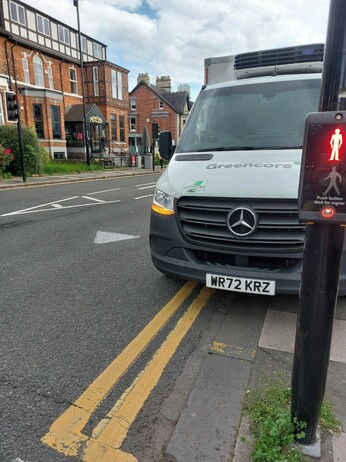 Delivery van parked on double yellow lines, partly on the pavement, right beside a signal controlled crossing, blocking the view of the road for pedestrians.
