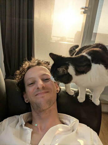 A white and brown cat sniffing the cheek of a human. That human is laying on the couch while the cat is sitting on one of the arm rests.