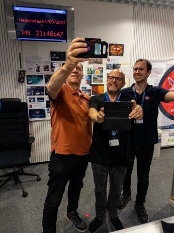 Three people standing facing the camera with mobile phones taking a picture of a digital mission clock on the wall behind them in a spacecraft control room at the European Space Operations Centre in Darmstadt.