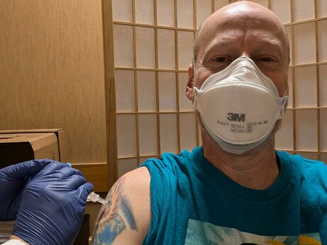 Selfie of me getting my COVID vaccination. I'm smiling under a mask, wearing a blue shirt with my sleeve rolled up. The pharmacists' hands are visible in purple gloves as he injects me.