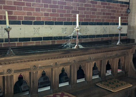 In the chancel of an old church is a carved wooden bench with a couple of  embroidered cushions on it. Behind it is a brick wall. 
Along the top of the bench is a line of three tall white candles in ornate candlesticks. A spider web joins the candles. Sunlight hits the centre candle, which is wreathed in cobweb. Both the candle and the web seem to glow.