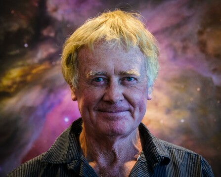 A headshot picture of John Hutchings standing in front of a large print of the Hubble Space Telescope mosaic of the Orion Nebula.