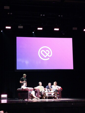 People seated on the stage on a couch recording a podcast. behind them the heart of clojure logo is projected