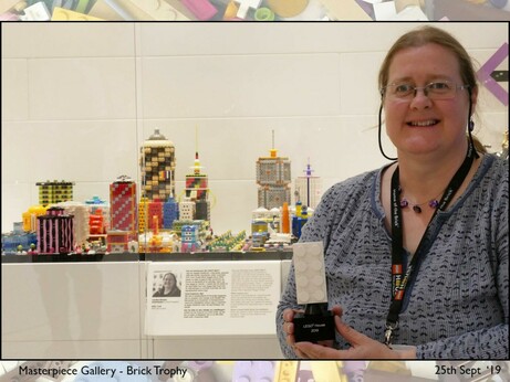 Me standing in front of my Micropolis MOCs in a brightly lit display cabinet, holding a white upscaled 2-4 brick trophy