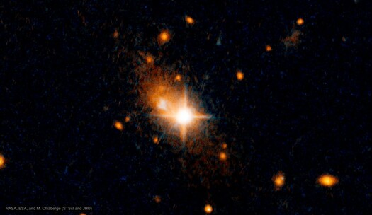 This image, taken by NASA's Hubble Space Telescope, reveals a runaway quasar fleeing from its galaxy's central hub. Near the center of the image is an elongated oval of orange light. Just to the right of the center inside the oval is a bright white circle with four diffraction spikes. That white circle is a quasar, which is a supermassive black hole that is actively pulling in material. The material produces the light we see, shining brightly as it is accelerated toward the black hole. But the quasar is not at the center of the galaxy. It has been jettisoned and is moving away. The rest of the image is dotted with a few orange spots. The image is watermarked “NASA, ESA, and M. Chiaberge (STScI and JHU).”