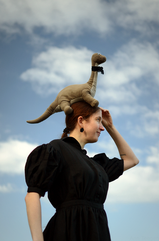 Foto von Ann mit ihrem niedlichen kleinen grauen Stoffbrontosaurier Gertrude auf dem Kopf, beide in die Ferne guckend vor einem blassblauen Himmel mit weißen Wolken. Ann trägt ein schwarzes verspieltes Leinenkleid und Gertrude eine schwarze Spitzenschleife.