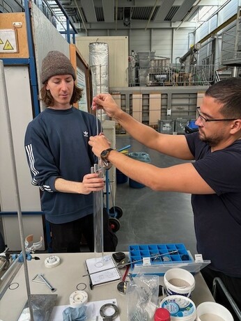Two individuals are working together in a laboratory setting. One person, wearing a beanie and a sweatshirt, is holding a long, transparent tube, while the other person, in a short-sleeved shirt and glasses, is adjusting a component connected to
