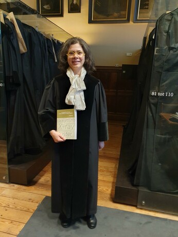 Me in a black university dress holding a PhD thesis in X-ray astronomy in a room with many more university togas hanging around.