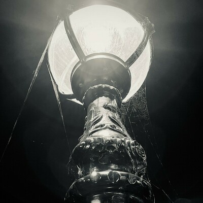 Square monochrome photo, looking up at a reasonably ornamental lamp outside Warrender Baths. At the top is a glowing orb, with cobwebs hanging from it. In the lower middle is a cast metal column, with partially lit ornamental patterns.