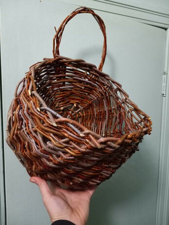 A hand holding up a woven willow basket, designed to hang on a wall, with a flat back and a rounded front, and a handle sticking out the top