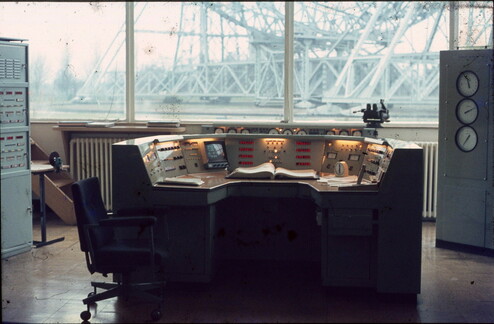 A window looks out onto the base of a huge radio telescope.  In the foreground is a control room, with a very classic control semicurcular control desk with many switches, a CRT, 7 seg displays and many switches.   And a mechanical stop watch.  To the sides are a rack of equipment and another cabinet with three large circular dials.