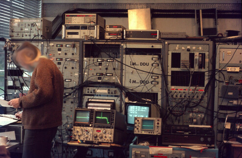 A person (whose face I've blurred) stands in front of a set of 6 racks containing all manner of switches, knobs and dials and displays.  In front of them are 3 oldish CRT scopes (including a classic Tek) and a CRT monitor with large white text.  More test equipment is piled on top of the cabinets.  Coax cables go everywhere.