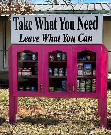 A vibrant pink community pantry with a sign on top that reads, "Take What You Need, Leave What You Can." The shelves are filled with various canned goods and pantry staples like pasta, soups, and beans. The phrase humorously aligns with the contents, as the pantry is filled with "cans," creating a playful unintended pun. The scene reflects generosity and community spirit in action.