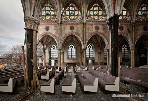 A side view of a decayed but ornate church. Rows of pews extend out ahead. The wall has been torn down on the left, revealing the sky