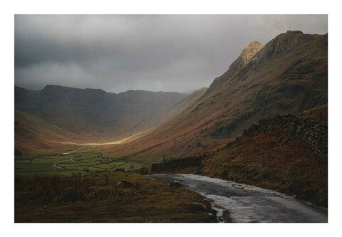 A photo of a Lake District scene. A road leads down with mountains on the right and a valley in sunlight below.