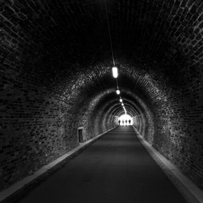 Four people approach the sunlit end of the tunnel. 

Roof lights along the roof of the tunnel provide sections of light and dark.