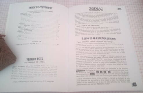 Foto de un ejemplar físico de la BETA preliminar de "Zodiac 2.5: System Reference Document". Abierto por la segunda y tercera página, donde se ven el índice de contenidos, una nota sobre el Beta-Testing, una breve descripción sobre el sistema Zodiac y unas indicaciones sobre "cómo usar este documento" que explican la convención sobre el uso de negrita, cursiva y otros recursos visuales que ayuden a mejorar la comprensión lectora.