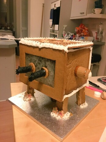 On a kitchen counter, on a tray, stands a gingerbread replica of a glovebox  used in chemistry labs. It has 4 legs supporting a literal box. Two gloves stick out from the center fron. It's trimmed with white frosting, silver candy spheres, and other edible decorations.