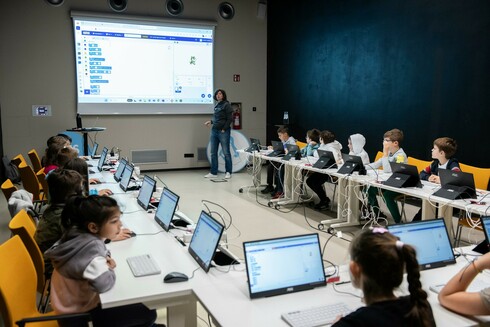 Many children sitting in a U table setup with computers. At the front wall a projector shows a screen with scratch running, trainer standing next to it.