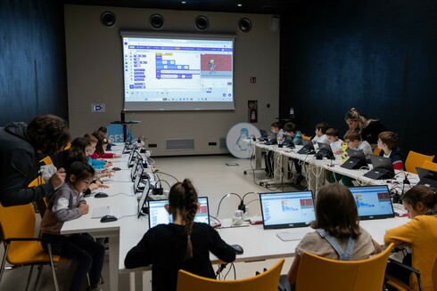 Many children sitting in a U table setup with computers all programming scratch. At the front wall a projector shows a screen with the source code of a computer program in scratch.