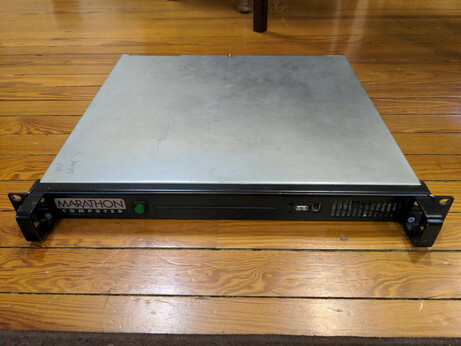 A 1U rackmount computer lying on a wood floor. The front and handles are painted black and the top of the case is bare metal. The Marathon Computer logotype is on the left, then a round green button, CD slot, USB A and FireWire 400 ports, and a grill on the right.