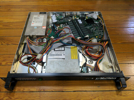 A 1U computer with the top removed, lying on a wood floor. A motherboard with RAM, PSU, and CD reader are visible. Power and ribbon cables lie loose in the open space next to the CD reader.