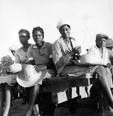 The image depicts four African American women sitting on a bench or cart, dressed in casual clothing and wearing wide-brimmed hats. They appear to be waiting for something or someone, as their expressions are neutral and contemplative.

In the background is another person who seems to be part of the group but is partially obscured from view by others. The setting appears rural with open skies suggesting an outdoor environment during daylight hours. There's a sense of stillness in the image, captured without any visible movement or action taking place at this moment.

Overall, the photograph conveys a historical context and tells a story about these women possibly being transported to another location for labor-related reasons as part of the Great Migration from rural southern states seeking better economic opportunities. The black-and-white nature of the photo indicates that it is an old image, likely taken during or before World War II.

The caption provided suggests this photograph was associated with "Women being transported from Memphis, Tennessee to an Arkansas plantation," indicating a historical context related to migration and labor in America's southern states during those times. The specific dates mentioned are July 1937; however, without additional metadata information about the image itself (such as EXIF data), it is not possible for me to confirm this with certainty based on visual analysis alone.

This phot [...]