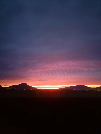 A breathtaking sunset over a serene landscape with silhouetted mountains in the distance. The sky is an ethereal gradient of deep blue at the top, transitioning into vivid shades of magenta, pink, and fiery orange near the horizon. Tiny city lights twinkle across the bottom of the frame, adding a touch of warmth to the tranquil scene.