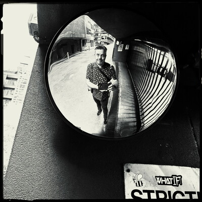 A black and white photo of myself in the reflection of a curved mirror at a car park entrance. I’m wearing a dark shirt, and a camera hangs at my side. A sign at the bottom right of the frame shows the trip of the word “STRICT”.