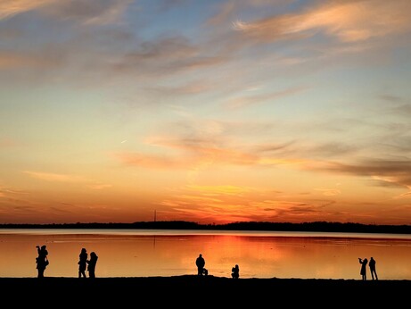 Sonnenuntergang an einem See. Himmel und See strahlen in rotorange Tönen. Im Vordergrund die Silhouetten einiger Menschen am Strand.