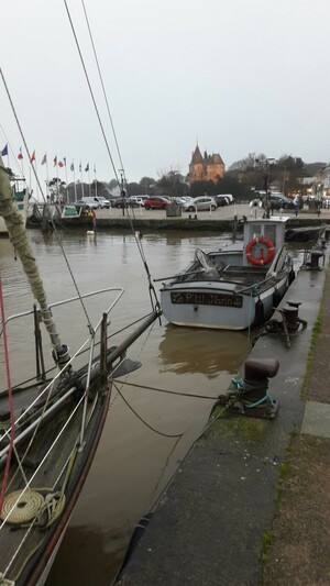 Une vue du port de Pornic au crépuscule par temps gris, à l'horizon le château aux tons orange de son système d'illumination. À gauche, un alignement de drapeaux en bord du quai rive droite. En premier plan, un cordage bien lové à l'avant du pont en bois d'un voilier à quai.