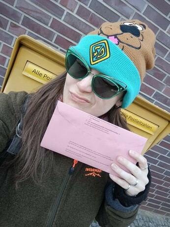 A photo of myself in front of a postbox, holding up my mail ballot. The yellow postbox is stood in front of a brick wall, I'm wearing my Scooby Doo hat, green sunglasses and a brown jacket while pouting my lips and winking.